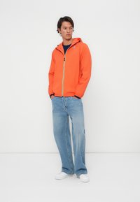 Chaqueta ligera naranja con capucha, cremallera en contraste y detalles en azul marino, combinada con jeans de corte relajado en azul claro y zapatillas blancas.