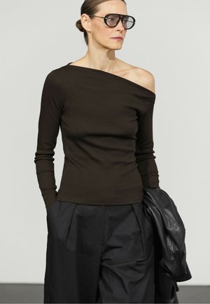 Femme portant de grandes lunettes de soleil, un haut noir à manches longues et épaules dénudées, un pantalon noir, tenant une veste en cuir noire.