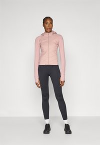 Világos rózsaszín, steppelt sportdzseki kapucnival, fekete leggings-szel párosítva. Hosszú ujjú, testhezálló kialakítású, sportcipővel viselve.