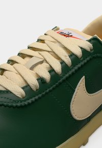 Σκούρο πράσινο δερμάτινο αθλητικό παπούτσι με μπεζ κορδόνια και λογότυπο swoosh, κοντινή λήψη του εμπρός δεσίματος και ραμμένες λεπτομέρειες στην πλευρική επιφάνεια.