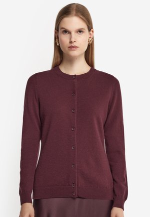 Donna che indossa un cardigan lungo a maniche lunghe, con bottoni, di colore prugna scuro e orecchini a cerchio dorati, su sfondo bianco semplice.