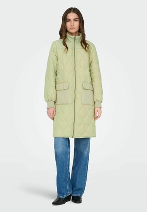 Femme portant une longue veste matelassée vert clair avec poches avant, jean bleu et chaussures noires à bout pointu, debout devant un fond blanc.