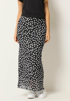 Frau trägt einen langen schwarzen Rock mit weißen Punkten, kombiniert mit weißen Turnschuhen und einem schwarzen Kurzarmtop, steht vor einer schlichten Wand.