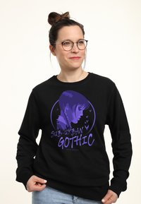Una donna con occhiali e capelli raccolti in uno chignon indossa una felpa nera con una grafica viola raffigurante il profilo laterale di una ragazza e la scritta "Sub-Urban Gothic".