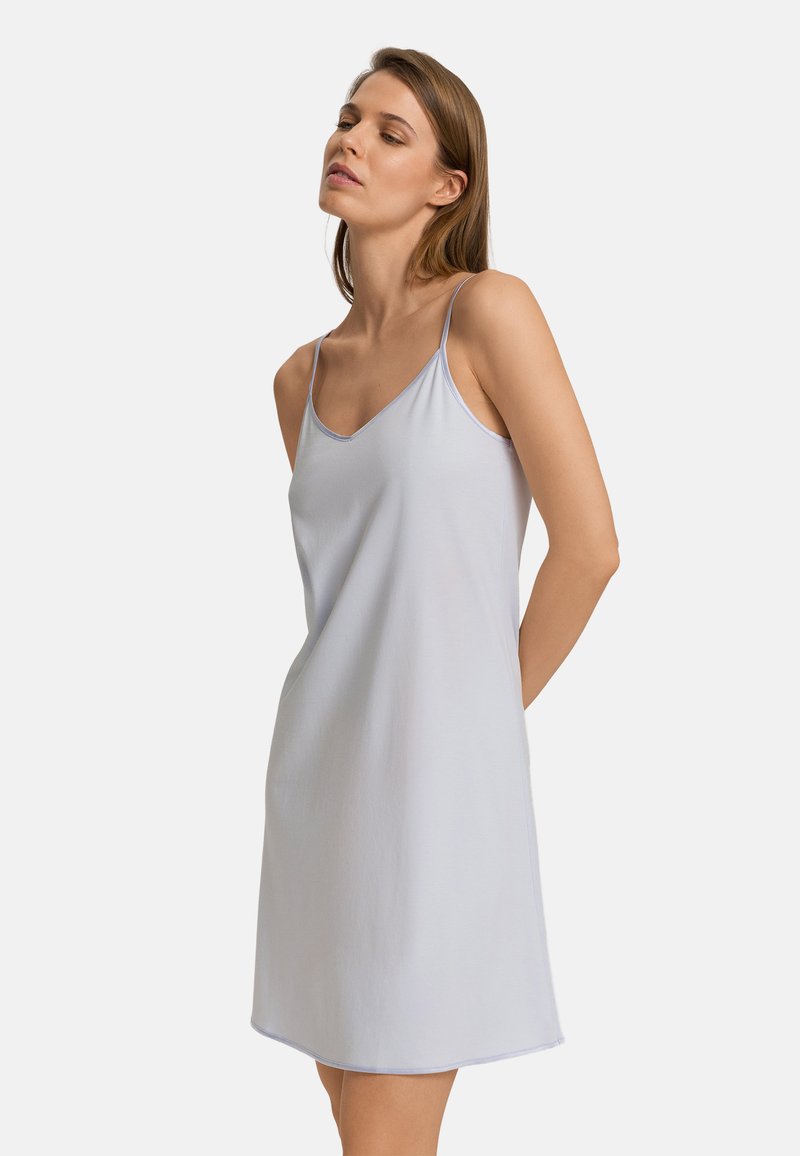 Nainen seisoo käsivarret selän takana, yllään hihaton, vaaleanharmaa liivimekko, jossa on ohuet olkaimet, valkoisella taustalla.