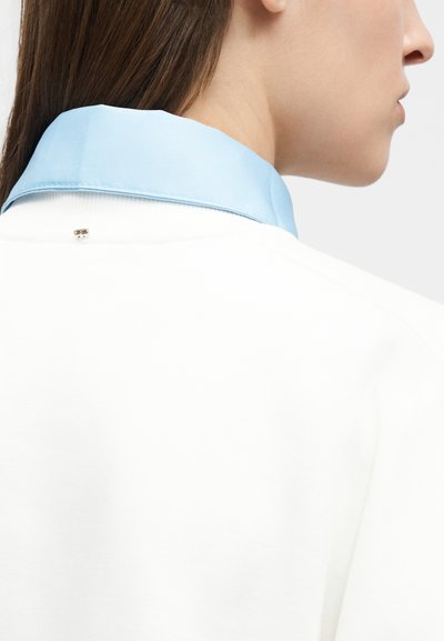 Προφίλ γυναίκας με ίσια καστανά μαλλιά, που φοράει λευκό ένδυμα με ανοιχτό γαλάζιο γιακά και μικρή μεταλλική λεπτομέρεια στο πίσω μέρος.