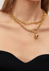Collier chaîne double couche dorée avec un pendentif rond porté autour du cou d'une femme au-dessus d'un top noir sans bretelles.