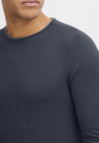 Maglione grigio scuro a maniche lunghe con scollatura a girocollo. Il tessuto è morbido, con un leggero rigato lungo il colletto e dettagli di cucitura sulle maniche.
