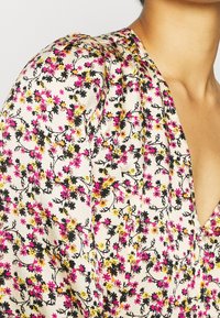 Femme portant un chemisier crème avec des manches bouffantes et un motif floral en rose, jaune et noir, doté d'un décolleté en V.