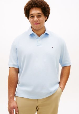 Jonge man met krullend haar en sproeten, gekleed in een lichtblauw poloshirt en beige broek, staand met één hand in zijn zak.