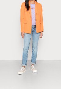 Woman wearing a bright orange blazer, light purple shirt, rolled-up light blue jeans, and beige sneakers standing on a plain light floor.