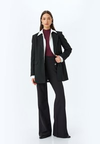 Zwart oversized blazer met witte kraag, gecombineerd met een getailleerde bordeauxrode top en uitlopende zwarte broek. Opvallende knoopdetail op de broek.
