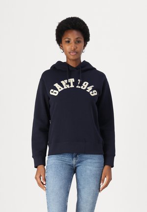Marine hettegenser laget av bomullsblanding, med "GANT 1949" logo i kremfarge, løs passform, ribbede mansjetter og nederkant. Kombinert med lyseblå jeans.