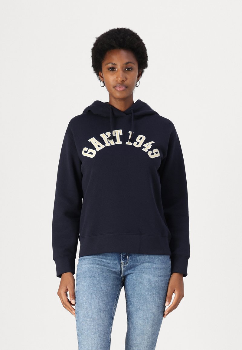 Sweatshirt à capuche navy en mélange de coton, arborant le logo "GANT 1949" en crème, coupe décontractée, poignets et ourlet côtelés. Assorti à un jean bleu clair.