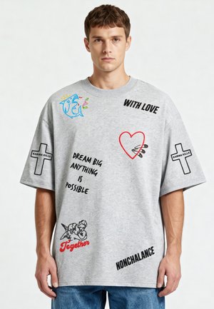 Jeune homme portant un T-shirt gris oversize avec des phrases, des croix étiquetées « Karma Kills », un cœur rouge, des chérubins et un graphisme de requin bleu.