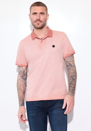 Mężczyzna o krótkich włosach i wytatuowanych ramionach, ubrany w różową koszulkę polo i ciemnoniebieskie dżinsy na białym tle.