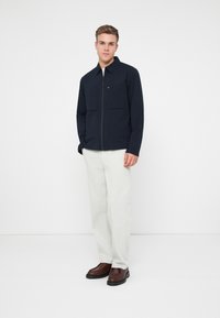 Ναυτικό μπουφάν με φερμουάρ και δύο μπροστινές τσέπες, συνδυασμένο με ανοιχτό γκρί παντελόνι και καφέ εσπαντρίγιες. Λείο ύφασμα με ελάχιστες λεπτομέρειες.