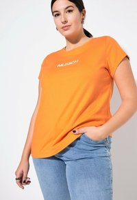 Orange Baumwoll-T-Shirt mit kurzen Ärmeln und gesticktem "MUNICH"-Logo auf der Brust, kombiniert mit hellblauen Jeans.