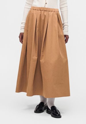 Person som bär en beige plisserad midikjol, vit långärmad topp, vita strumpor och svarta glansiga loafers, stående mot en enfärgad bakgrund.