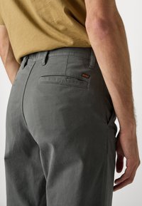 Homme portant un pantalon gris avec une poche arrière et une étiquette "BOSS", associé à une chemise de couleur moutarde, se tenant devant un fond uni.