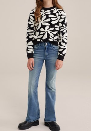 Jonge persoon met een zwart-wit gebloemde trui, vervaagde blauwe flare-jeans en zwarte chunky laarzen tegen een effen achtergrond.