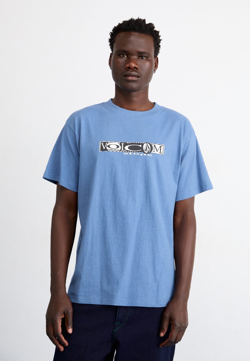 Tricou din bumbac de culoare albastră deschisă, cu mâneci scurte și guler rotund; are un logo grafic alb-negru în centru.
