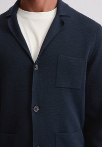 Marineblaue Strickjacke aus strukturiertem Strickstoff, mit Reverskragen, fünf Knöpfen und einer Brusttasche.