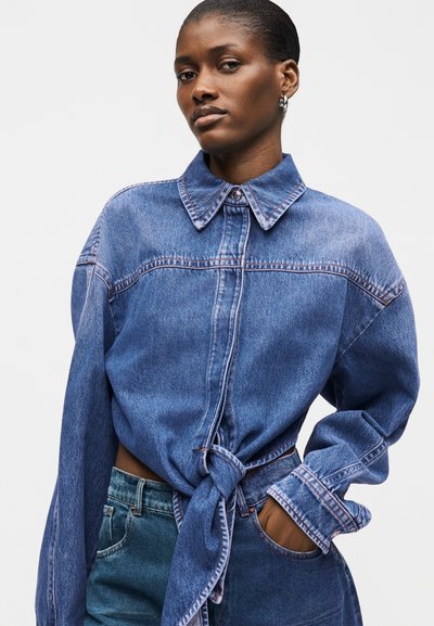 Γυναίκα που φοράει υπερμεγέθη μπλε τζιν πουκάμισο δεμένο στη μέση και τζιν παντελόνι του ίδιου χρώματος, ποζάροντας με το ένα χέρι στην τσέπη μπροστά σε απλό φόντο.