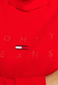 Sudadera de algodón roja con "TOMMY JEANS" bordado en dorado y un pequeño logotipo de bandera entre las palabras. Cuello alto y textura acanalada.