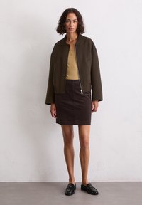 Veste bomber marron courte sur un débardeur beige, assortie à une jupe en corduroy marron foncé et des mocassins noirs. Détails simples de zip et de poches.