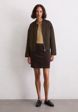 Veste bomber marron courte sur un débardeur beige, assortie à une jupe en corduroy marron foncé et des mocassins noirs. Détails simples de zip et de poches.