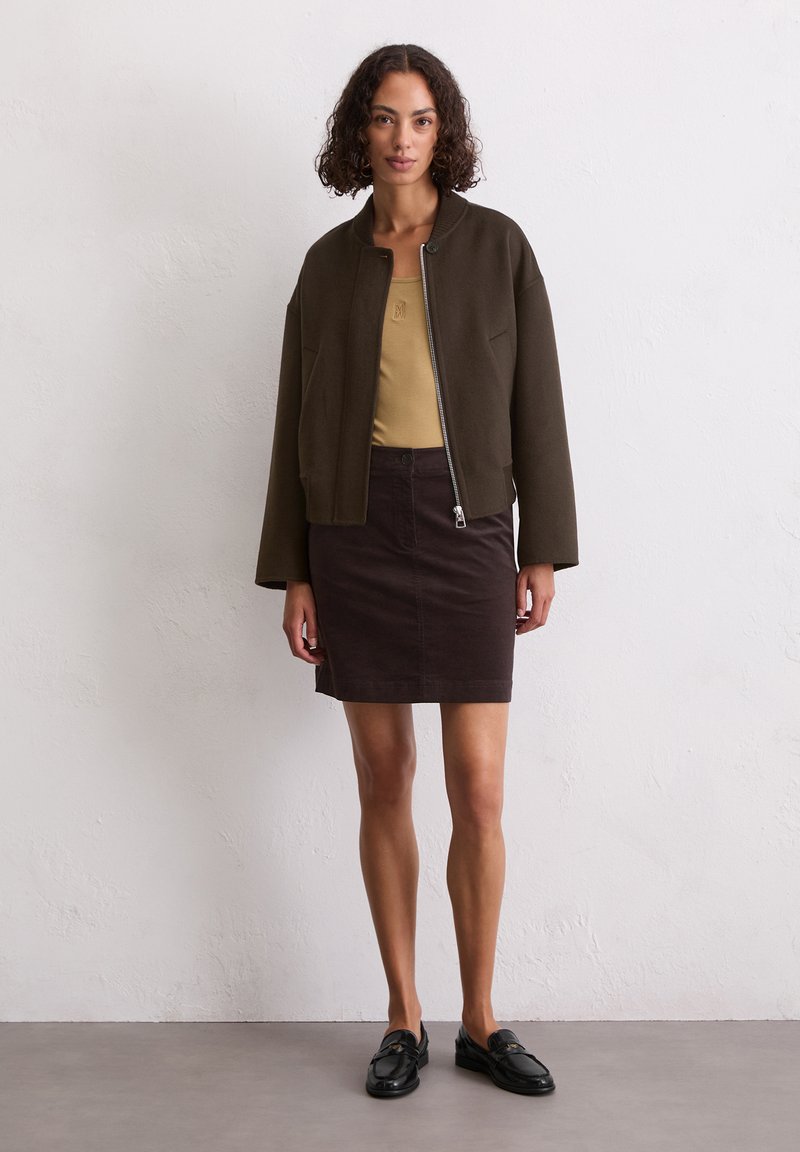 Veste bomber marron courte sur un débardeur beige, assortie à une jupe en corduroy marron foncé et des mocassins noirs. Détails simples de zip et de poches.