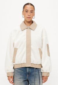 Witte fleecejas met beige accenten, voorzien van een knoopsluiting aan de voorkant, zijzakken en een ritszak op de mouw. Zachte textuur en ontspannen pasvorm.