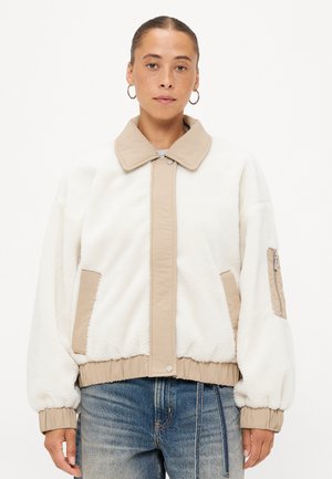 Witte fleecejas met beige accenten, voorzien van een knoopsluiting aan de voorkant, zijzakken en een ritszak op de mouw. Zachte textuur en ontspannen pasvorm.
