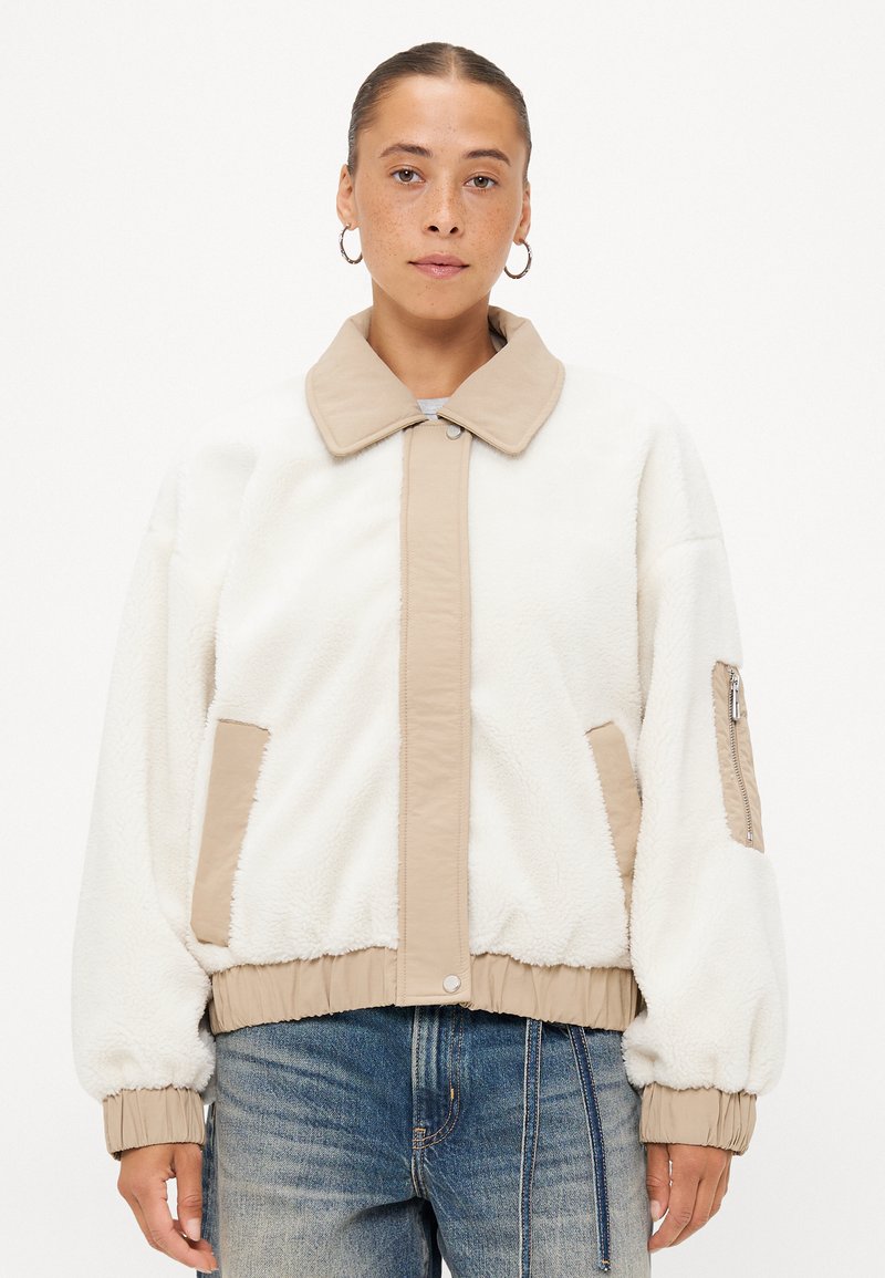 Witte fleecejas met beige accenten, voorzien van een knoopsluiting aan de voorkant, zijzakken en een ritszak op de mouw. Zachte textuur en ontspannen pasvorm.