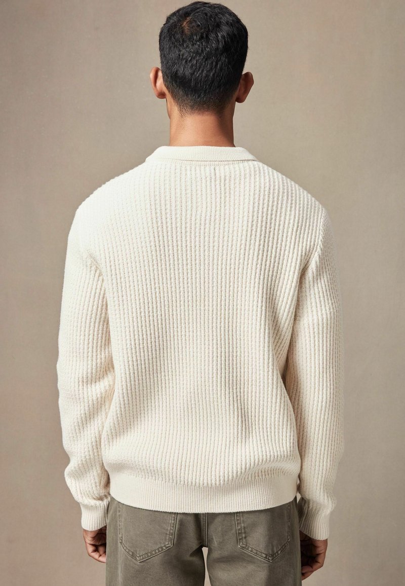 Crewneck-Pullover in Off-White, strukturiertem Rippenstrick; lange Ärmel mit eng anliegenden Bündchen, unverschönert, kombiniert mit olivgrünen Hosen.