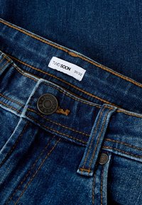 Tume sinises toonis teksapüksid vaseõmblustega. Neil on kahetooniline nööp ja valge kangasilt, millel on kiri "TWO SOON 31/32".