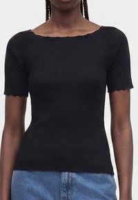 Femme portant un haut noir côtelé ajusté à manches courtes avec des bords festonnés, associé à un jean en denim bleu, sur un fond uni.