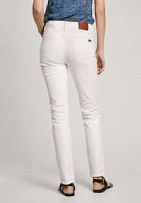 Hvite denimbukser med slim fit, med en lærlapp på bakliv og to baklommer. Sandaler synlige på føttene.