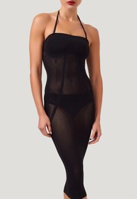 Donna che indossa un abito nero, trasparente, aderente con scollo all'americana sopra biancheria intima nera, mostrata dalle spalle fino a metà polpaccio su uno sfondo semplice.