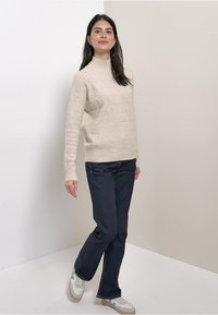 Pull en tricot beige avec un col montant et une texture côtelée, associé à un jean droit foncé et des baskets multicolores.
