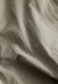 Vaaleanoliivinvihreä kangas, jossa on sileä, teksturoitu pinta. Kangas näyttää pehmeitä poimuja ja herkkiä ryppyjä, mikä luo hienovaraisia kiilto-eroja.