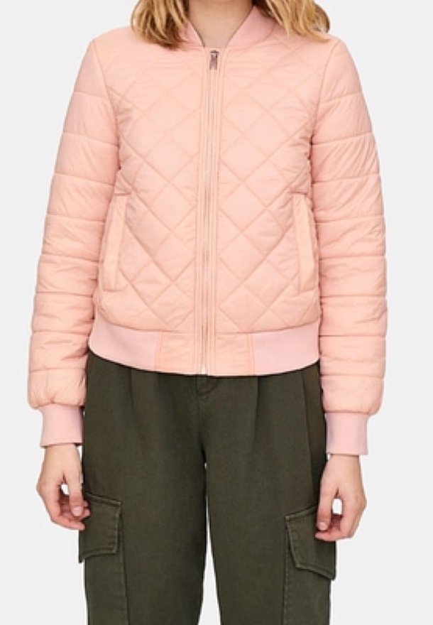 Personne portant une veste bomber matelassée rose clair avec une fermeture éclair à l'avant et un pantalon cargo vert avec des poches latérales.
