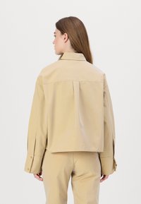 Beige oversized shirt in lightweight cotton with a back pleat. Features wide sleeves and button cuffs, styled with matching trousers.