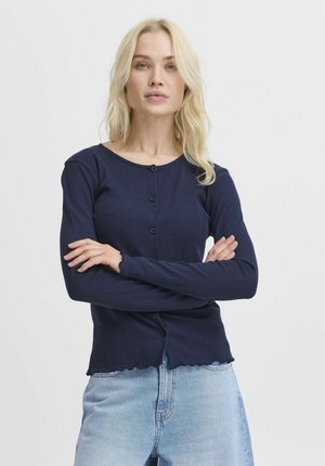 Námořnický pletený cardigan s dlouhými rukávy, kulatým výstřihem a pěti knoflíky. Materiál je měkký s vlnkovaným lemem, kombinovaný se světlým denimem.