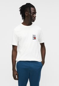 Λευκό βαμβακερό t-shirt με κοντά μανίκια, με μικρό λογότυπο κόκκινου, μπλε και μαύρου χρώματος στο αριστερό στήθος, συνδυασμένο με μπλε παντελόνια.