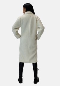 Lange crèmekleurige schapenvachtjas met kraag, rechte snit en zijzakken, gecombineerd met zwarte leren broek en enkellaarsjes.