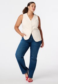 Beige mouwloos vest met knoopsluiting, gecombineerd met donkerblauwe spijkerbroek. Het model draagt rode hakken en gouden creolen en staat zelfverzekerd.