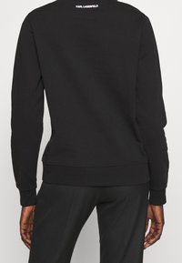 Sudadera negra con cuello redondo, mangas largas y puños acanalados. El logo "KARL LAGERFELD" está impreso en rosa en la parte superior de la espalda.