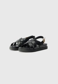 Black leather sandals with crossed straps and an adjustable ankle buckle. Flat sole with a textured finish. Simple, minimalist design.